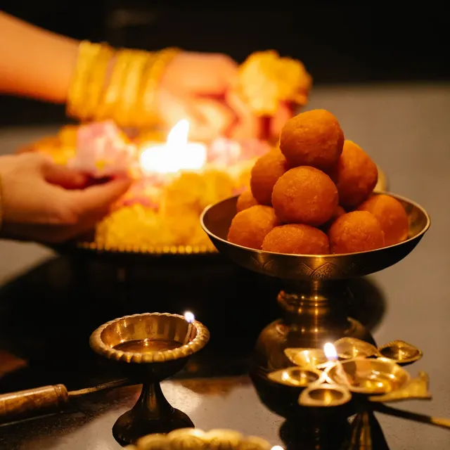 A brass bowl of laddoos beside lit brass diyas for Diwali puja